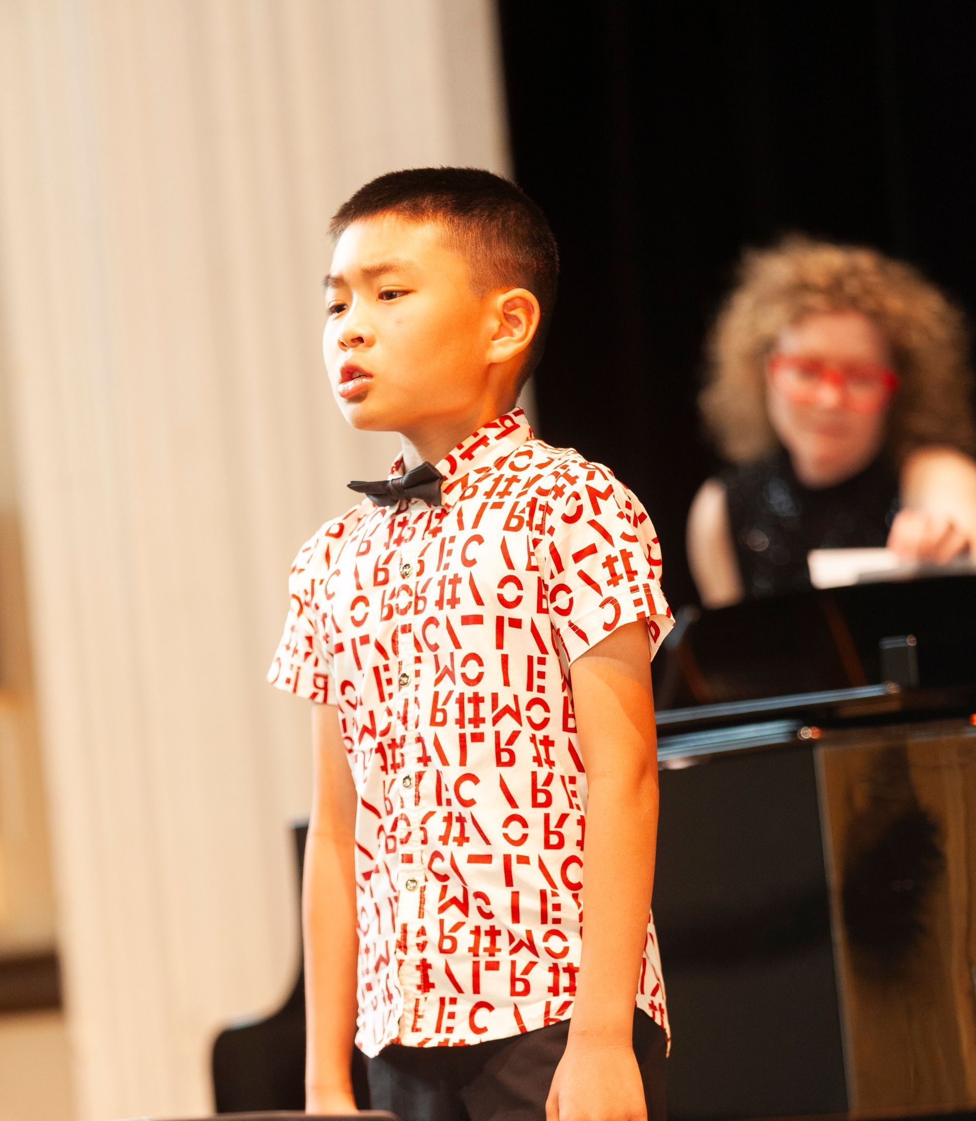 A young boy is standing in front of a piano wearing a shirt with letters on it
