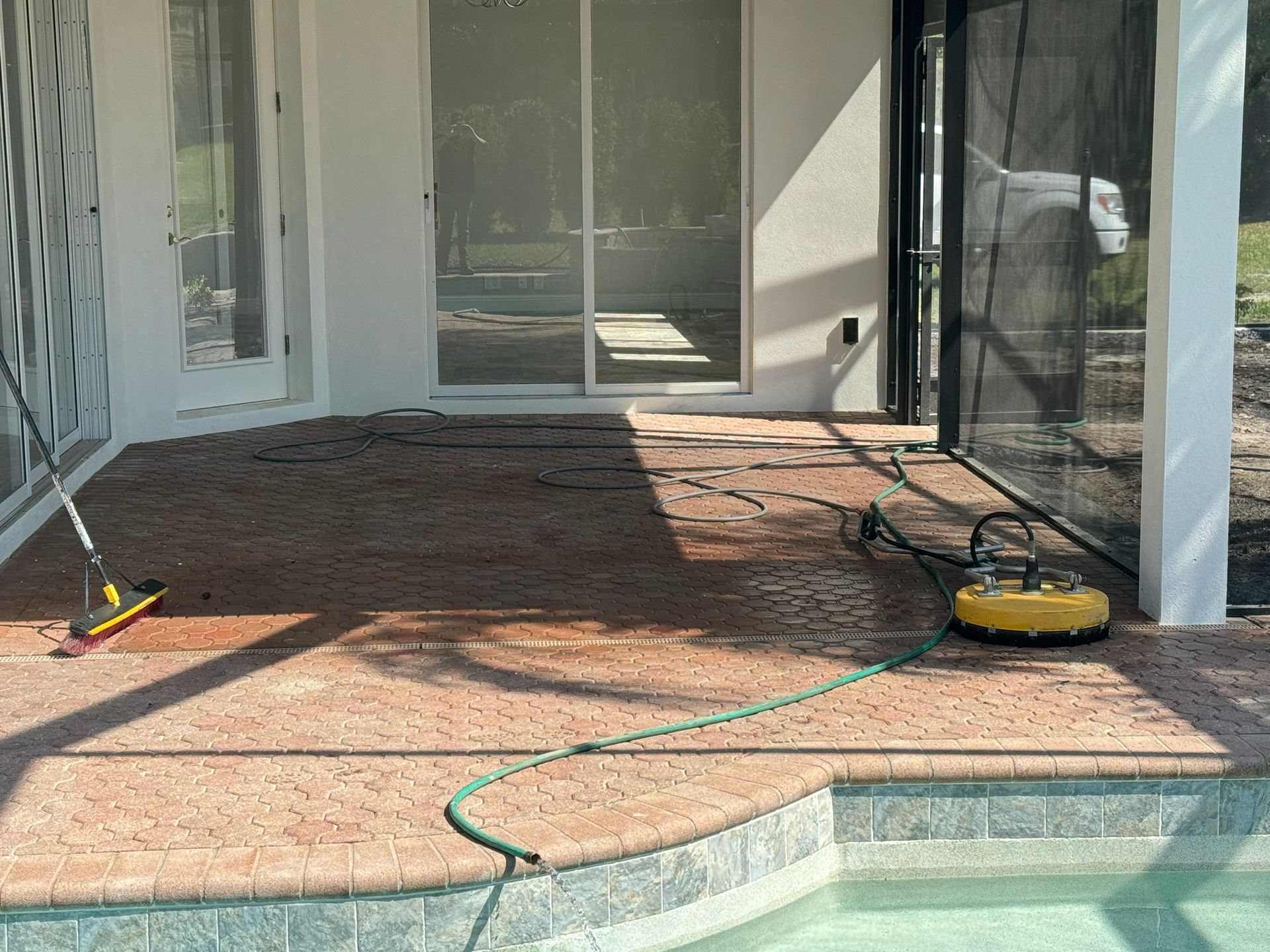 A patio surface cleaning tool and a broom sit on a brown stone pool deck next to a sliding glass door and screen enclosure.