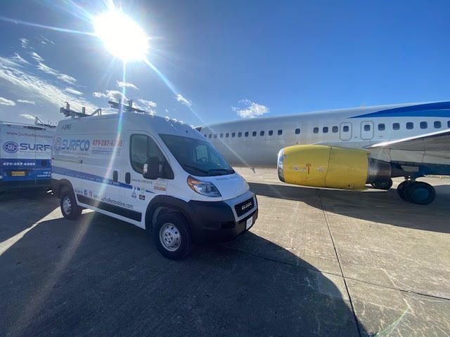 A white van is parked in front of an airplane.