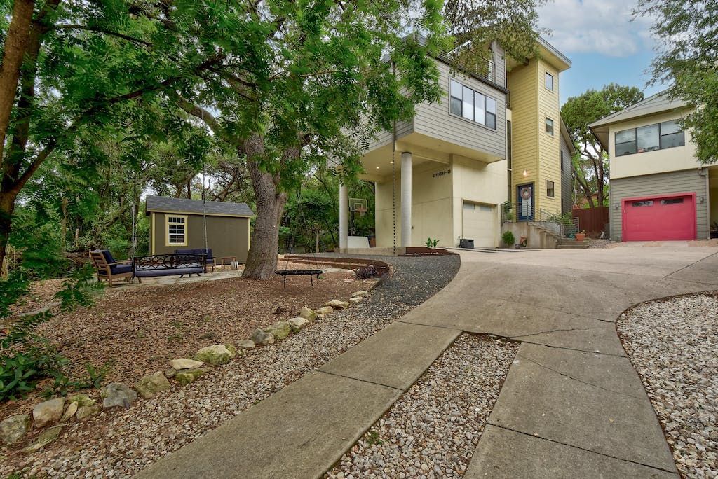 A house with a red garage door is surrounded by trees and gravel.