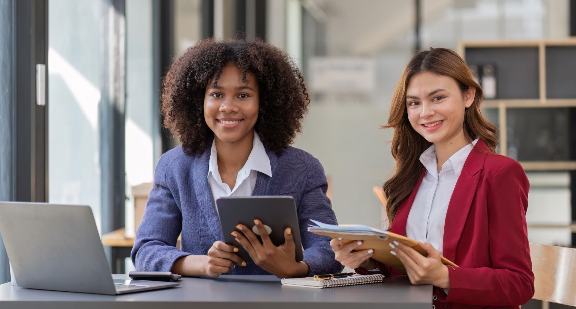 Two professionals smiling at a desk in an office, one holding a tablet and the other reviewing a document.
