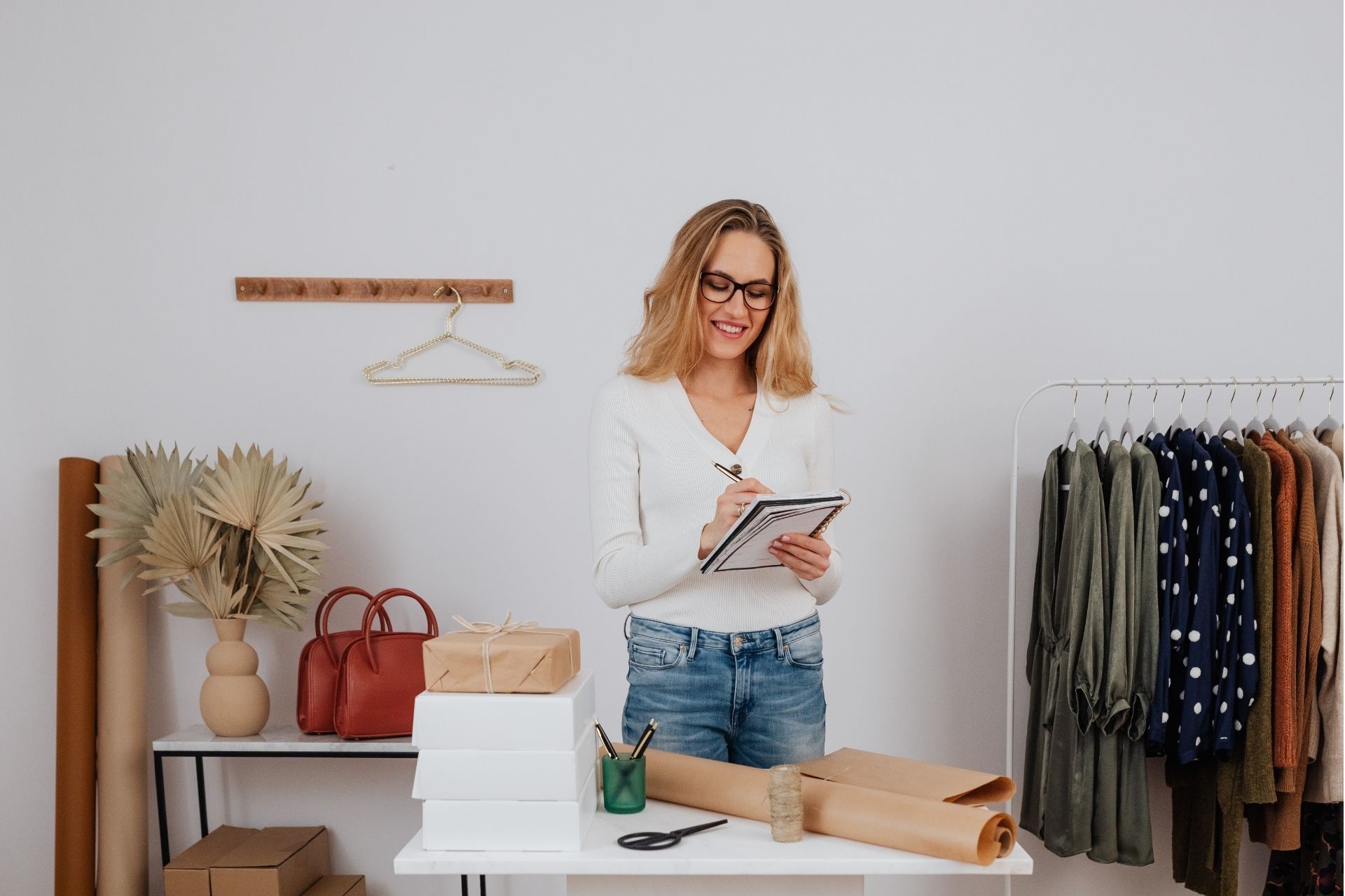 A person smiles while writing in a notebook at a table with wrapping supplies and a clothing rack in the background.