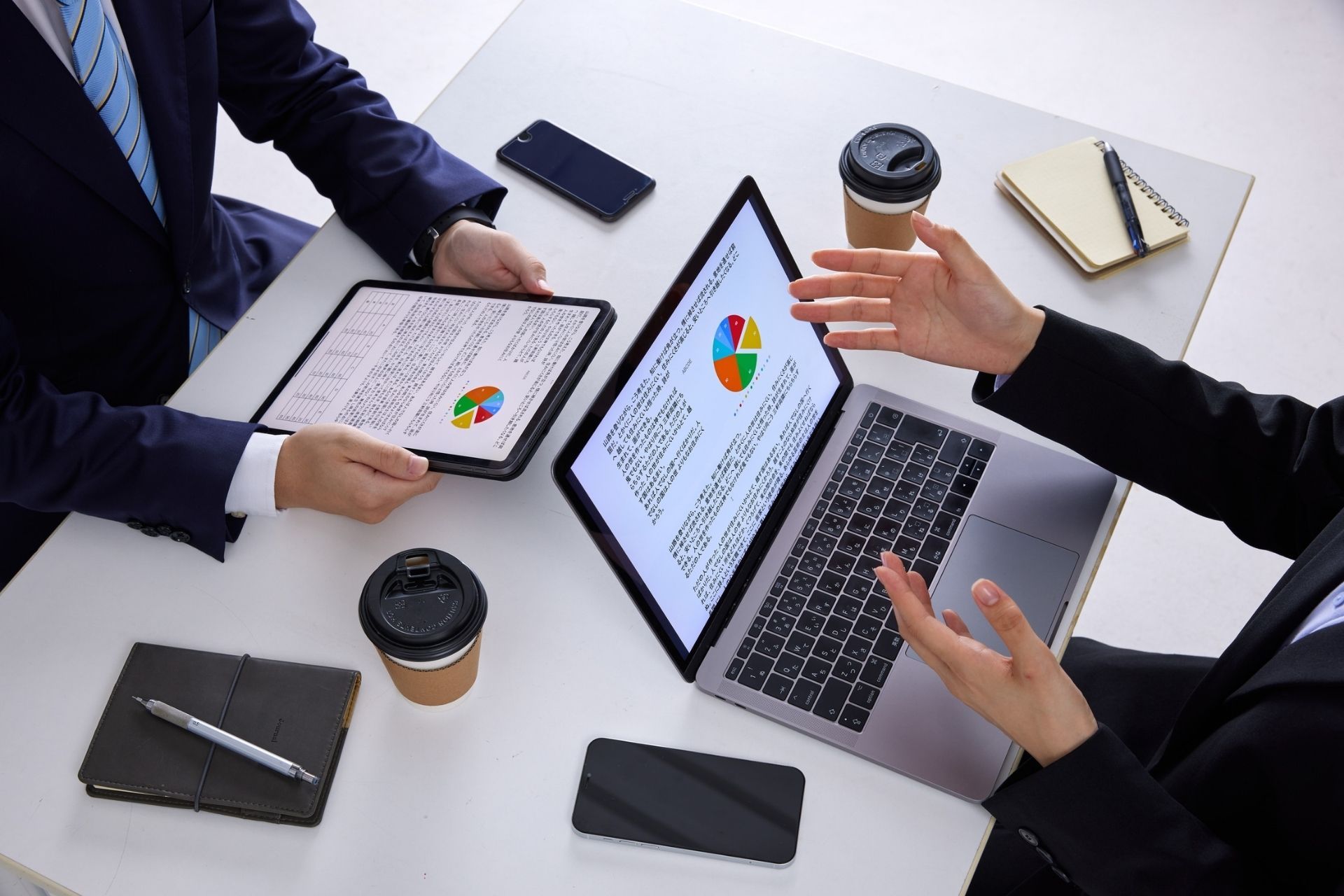 Two professionals in suits reviewing business data on a laptop and tablet at a white table with coffee and stationery.