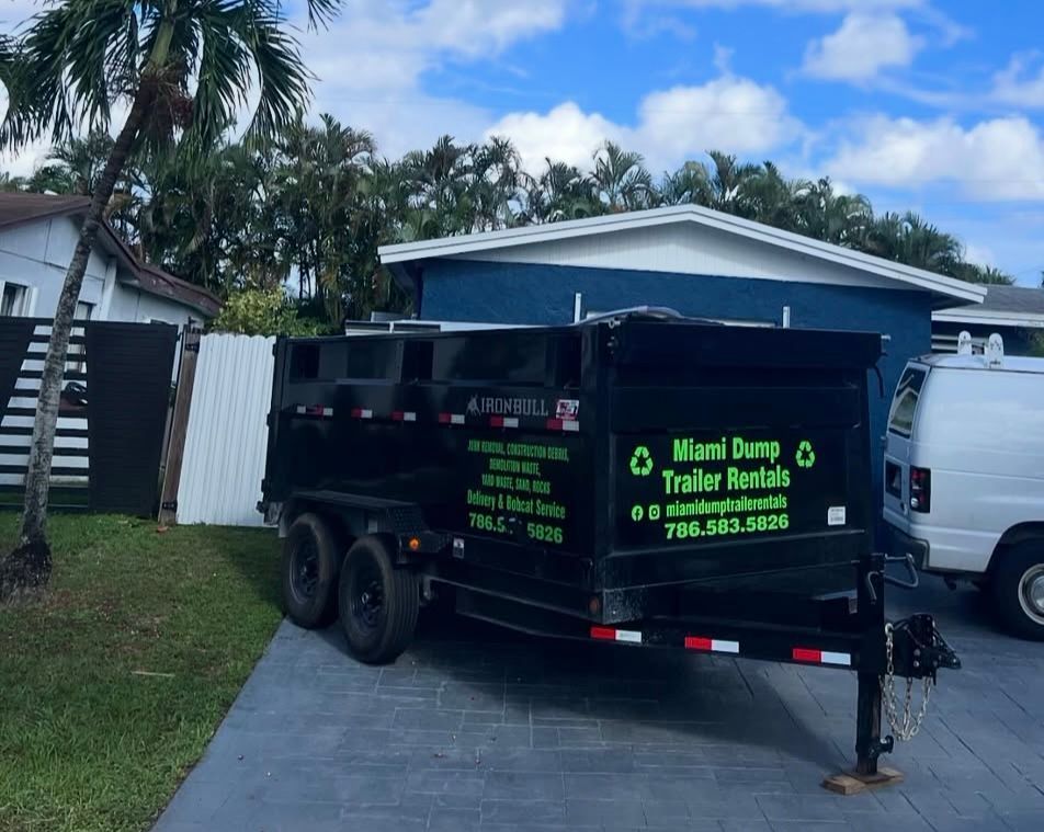 A black Miami Dump Trailer rental parked on a driveway next to a white house and a white van under a blue sky.