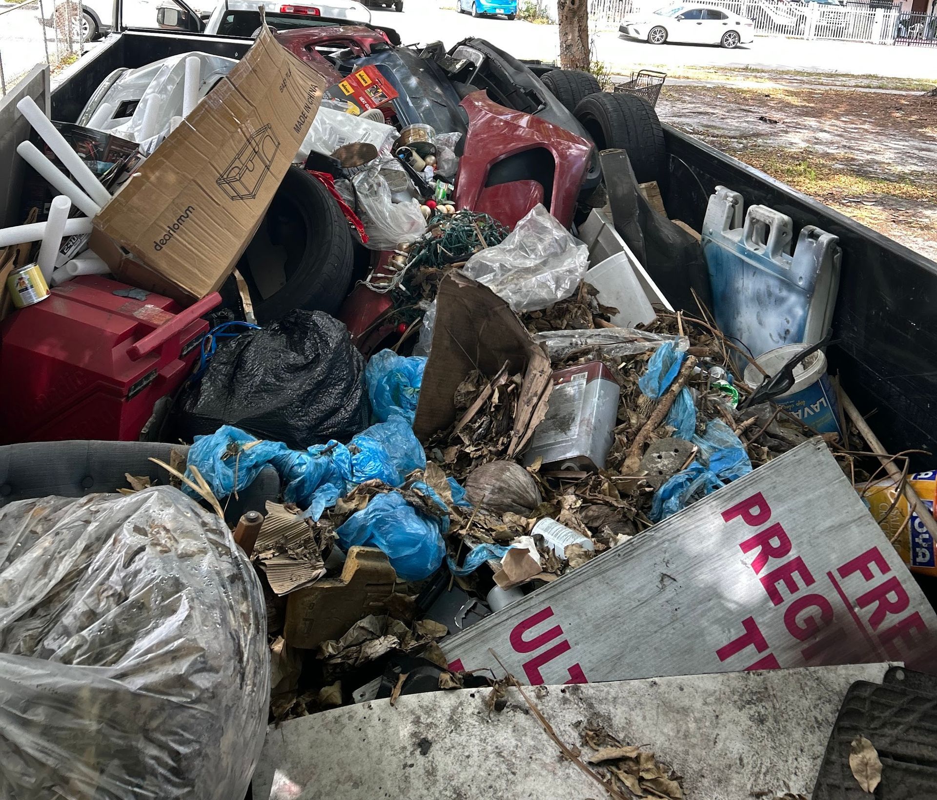A large metal dumpster filled with various debris, including cardboard boxes, car parts, tires, and discarded materials.