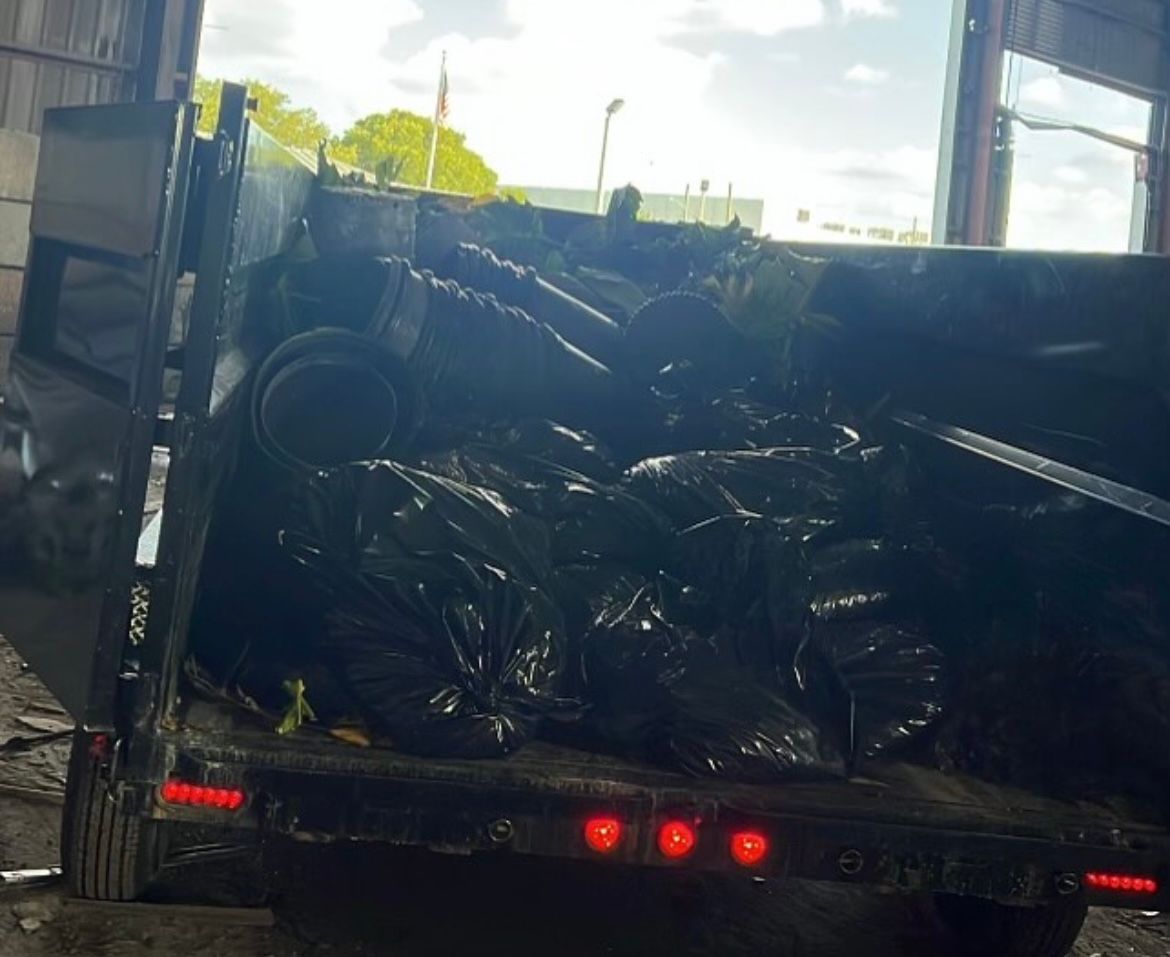 A dump trailer loaded with black garbage bags sits parked outdoors under a bright sky.