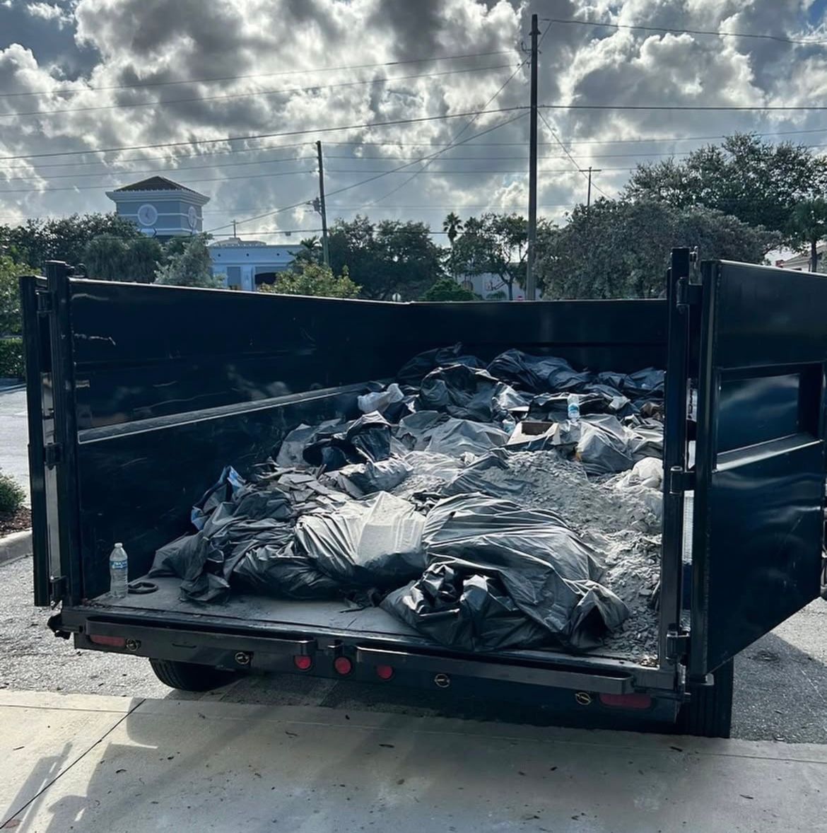 An open black roll-off dumpster filled with black trash bags and debris, set against a cloudy sky.