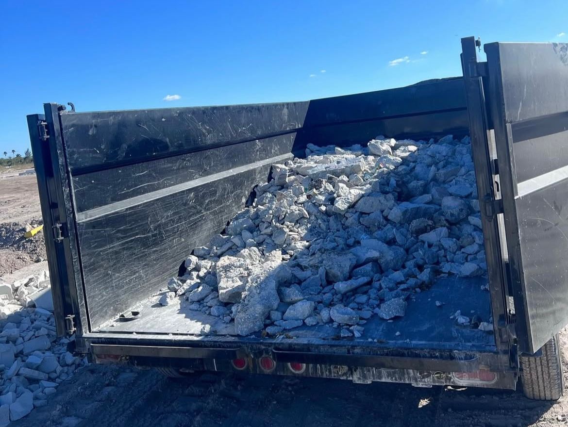 The back of a black dump trailer filled with large, light-colored rocks under a bright blue sky.