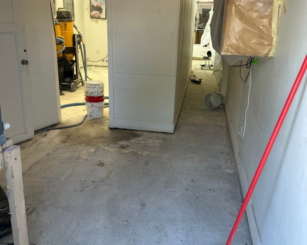 Concrete floor in a room under renovation with a vacuum cleaner, a bucket, and a red pole leaning against the wall.