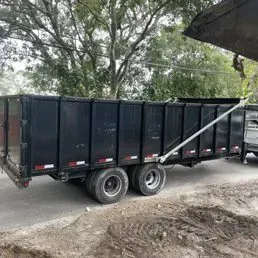 A large, black, rectangular dump trailer sits on a paved road next to trees and dirt.