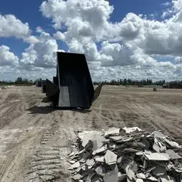 A metal dumpster sits in a large, open dirt lot near a pile of construction rubble under a cloudy, blue sky.