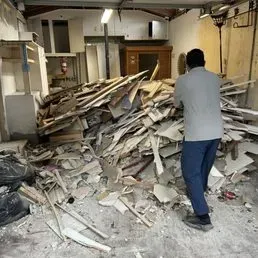 A person in a gray shirt and blue pants stands in a garage filled with construction debris and wooden scraps.