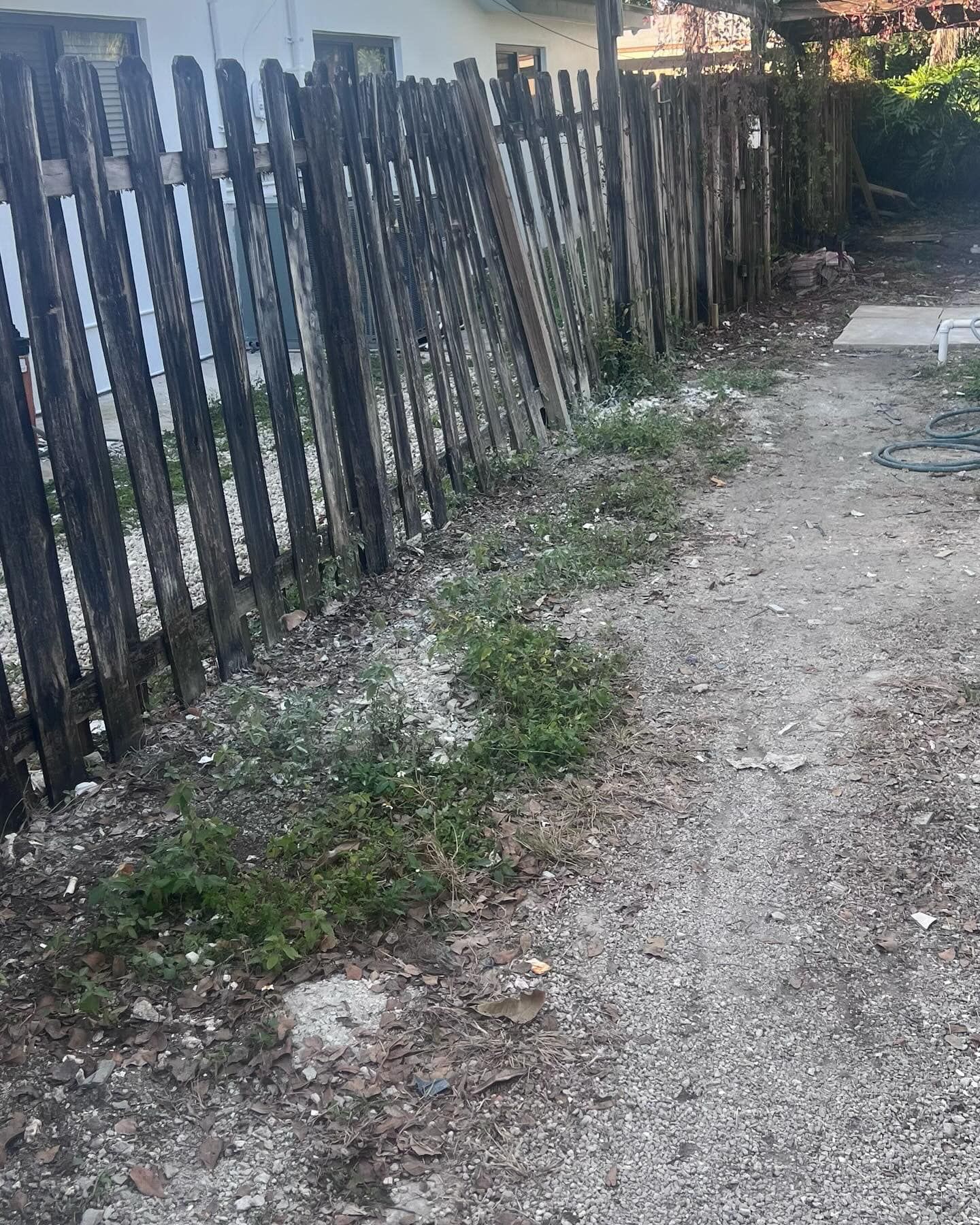 A weathered, dark wooden picket fence leans slightly over a gravel path with sparse patches of green weeds.