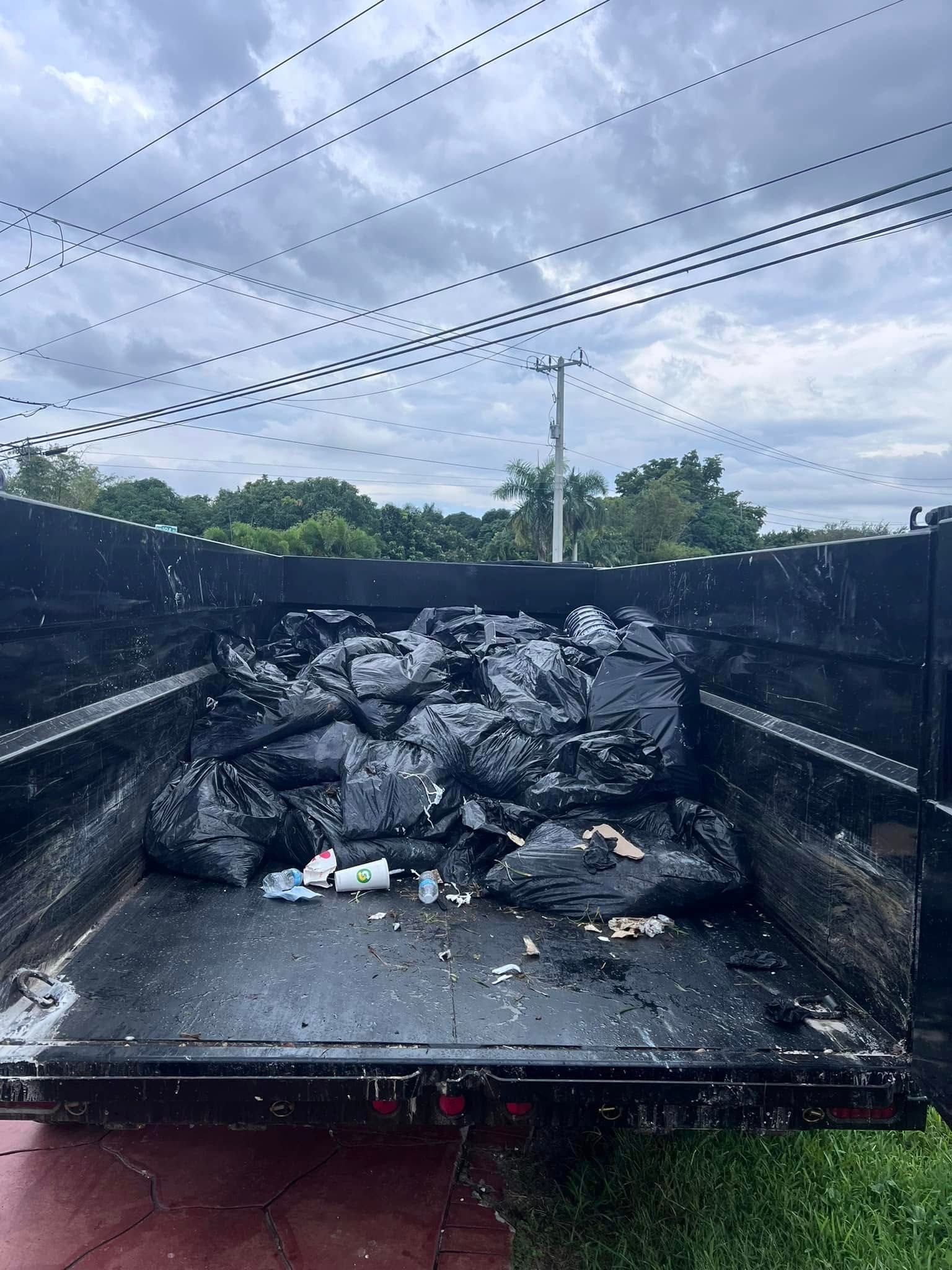 A black trailer filled with stacked, black garbage bags sits outdoors under a cloudy sky with utility lines above.