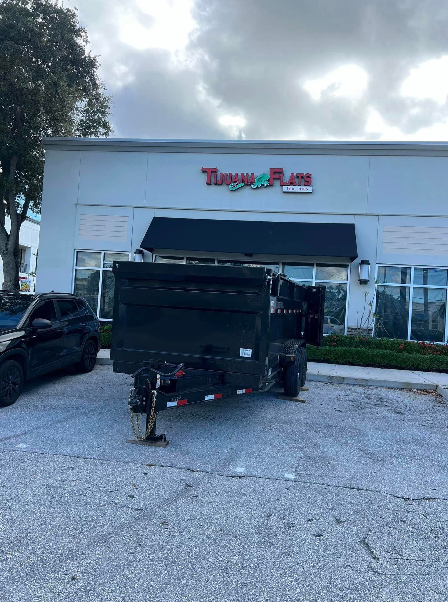 A black dump trailer parked on a gravel lot in front of a white building with the sign 