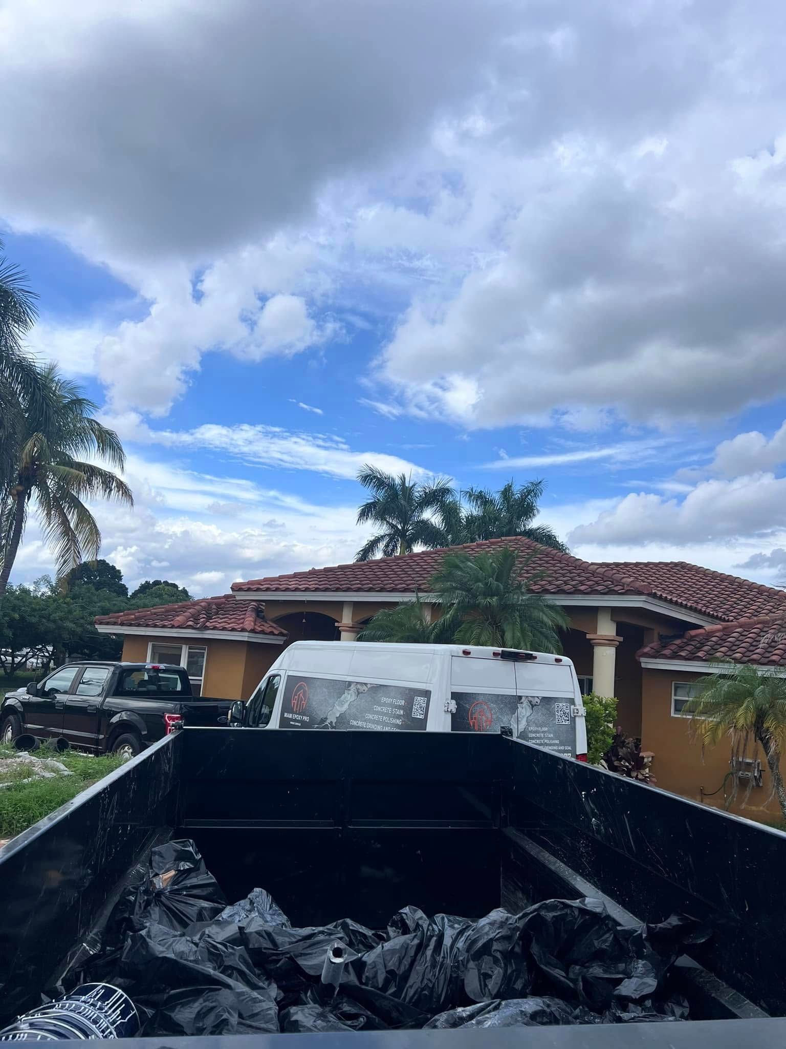 A black dumpster filled with dark bags sits in front of a residential house with a brown tile roof under a cloudy sky.