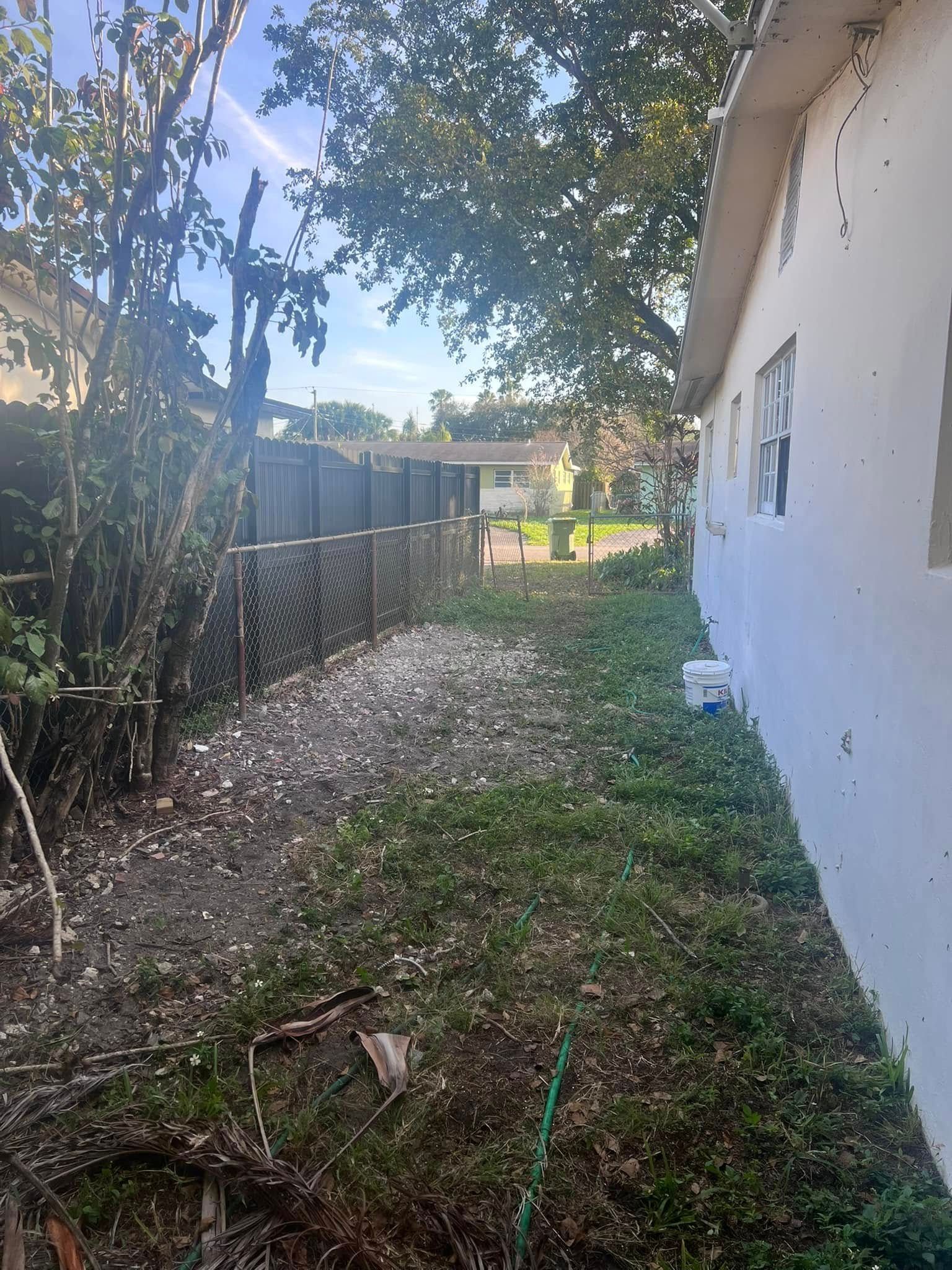 A narrow side yard between a white house wall and a dark metal fence, covered in patchy weeds, dirt, and light-colored rock.