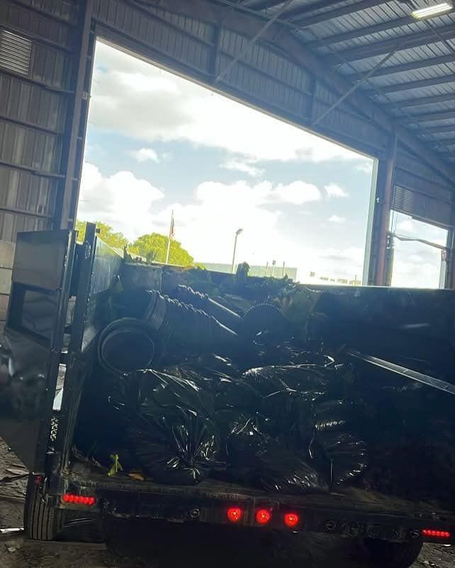 A trailer filled with black trash bags parked inside an open-air warehouse or bay with bright daylight in the background.