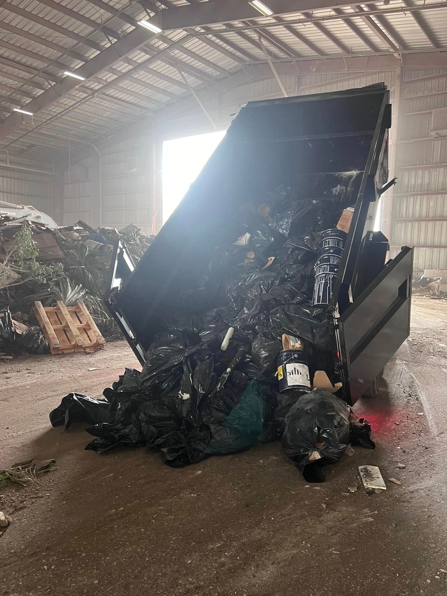A dark, industrial waste container tipped over inside a warehouse, spilling a pile of black debris onto the concrete floor.