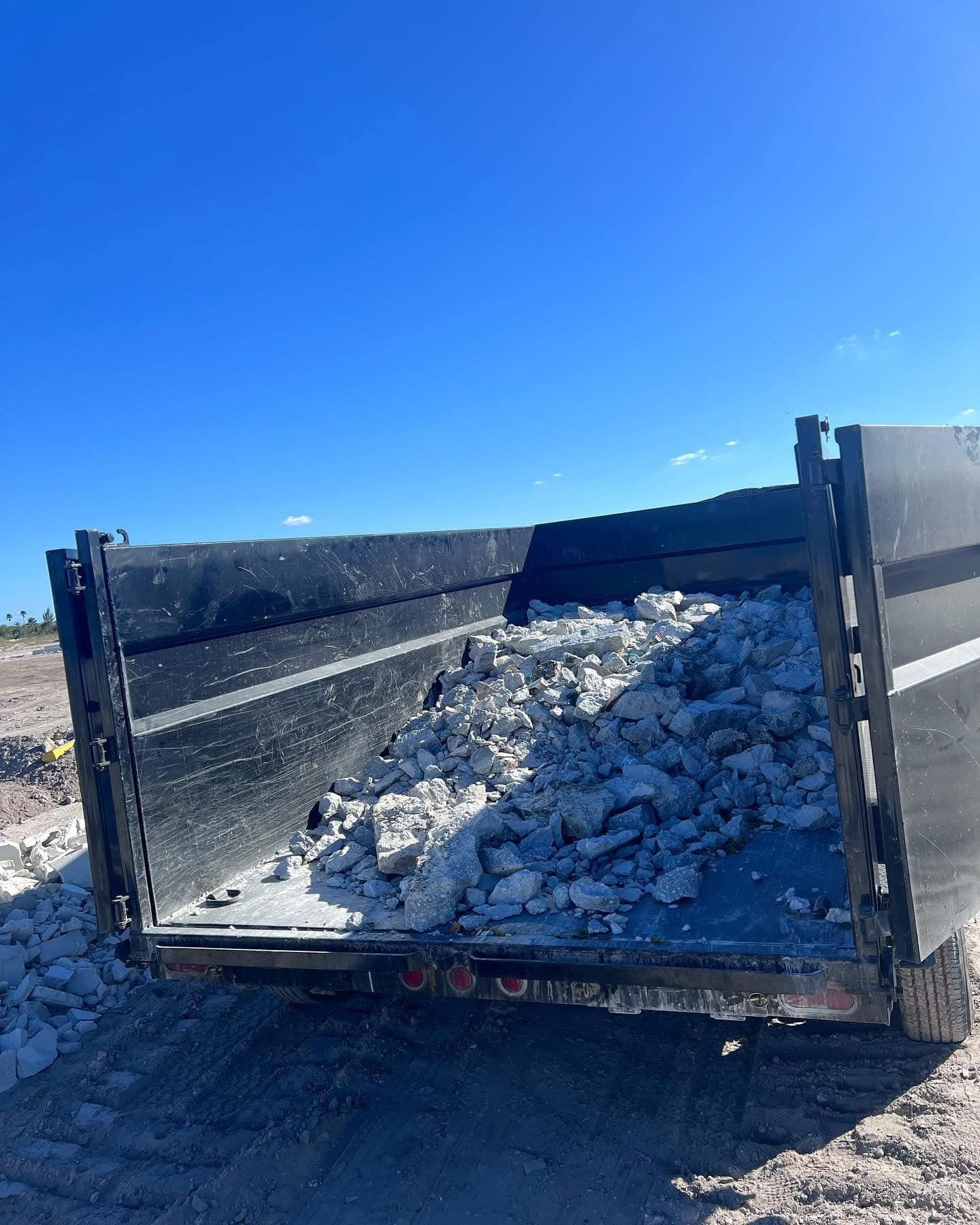 A black dump trailer bed partially filled with chunks of gray rock against a bright, clear blue sky.
