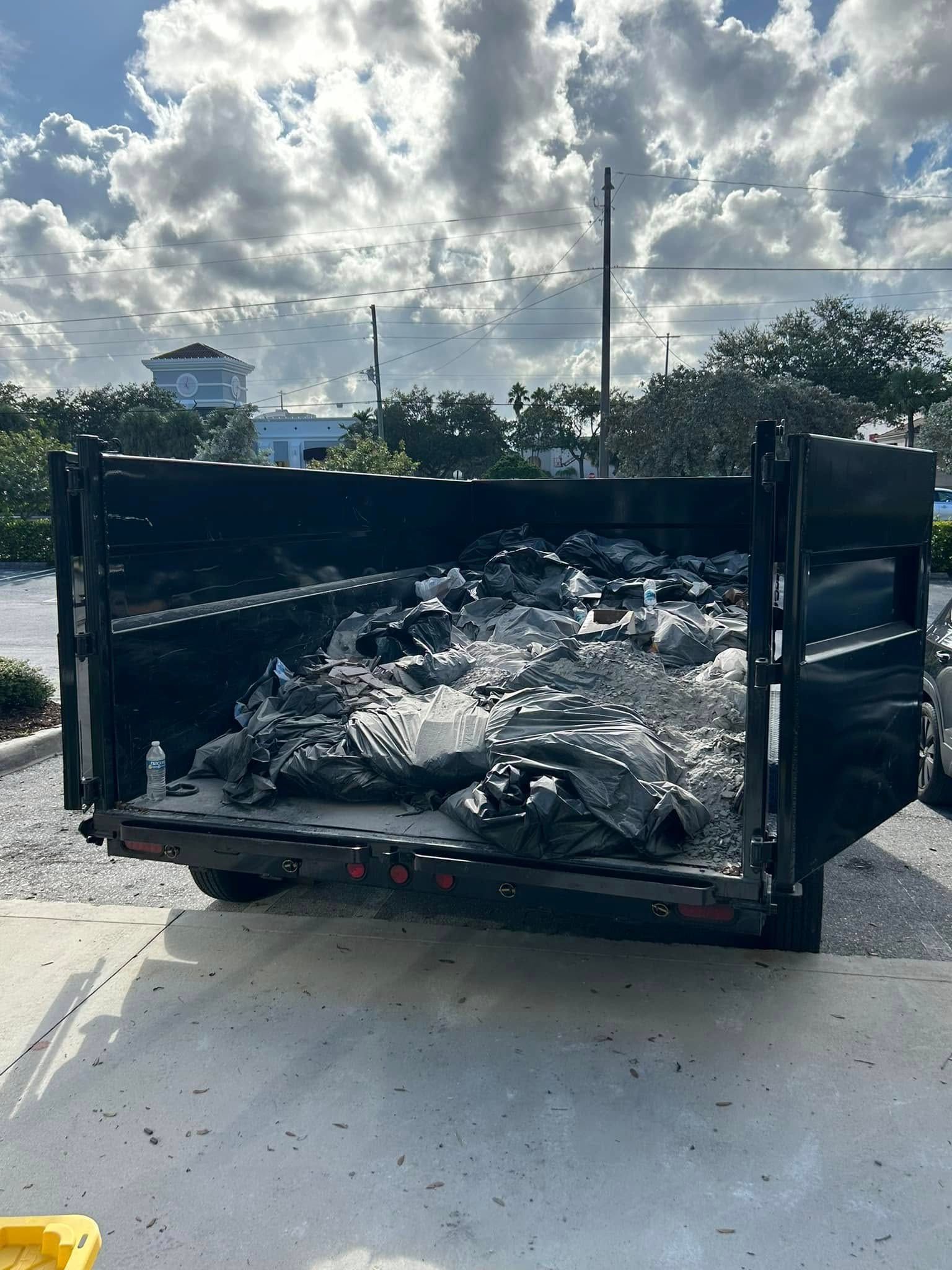 A black dumpster filled with various items and trash bags, parked on a concrete surface under a cloudy sky.