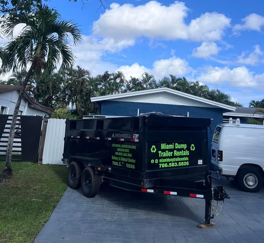A black dumpster trailer parked on a concrete driveway in front of a blue house under a cloudy sky.