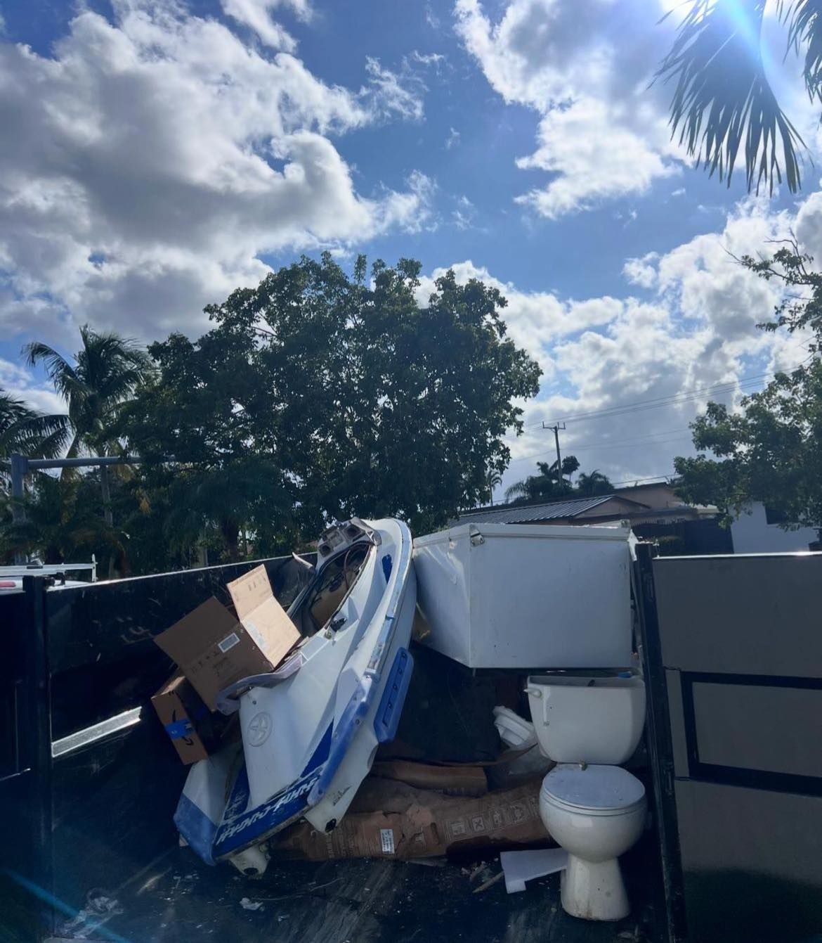 A white boat, a cardboard box, and a white toilet sit in a large metal dumpster under a blue, cloudy sky.