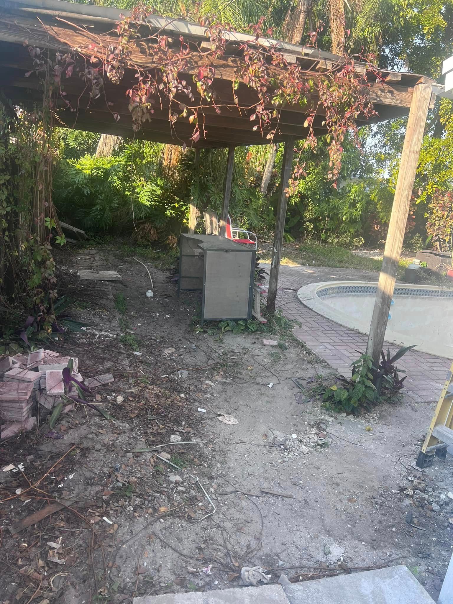 A wooden pergola covered in vines stands over a small outdoor bar area next to a pool deck with dirt and scattered debris.