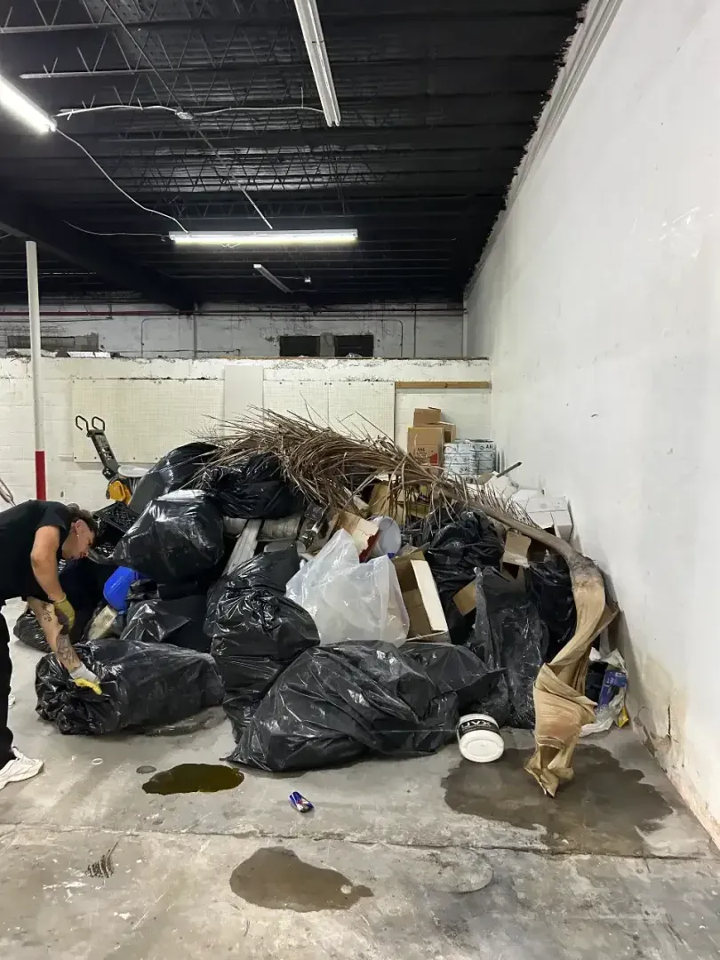 A person in a warehouse kneels beside a large, messy pile of trash bags, debris, and cardboard against a white wall.