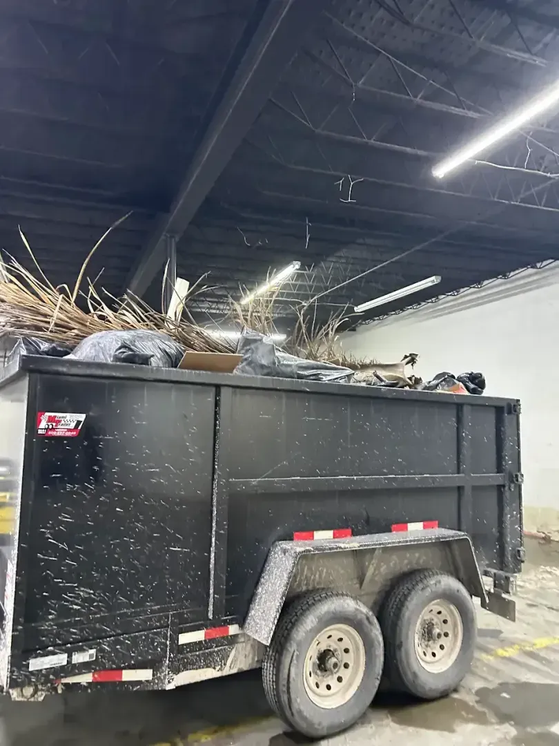 A black dump trailer filled with yard waste and debris, parked inside an indoor warehouse with overhead lighting.