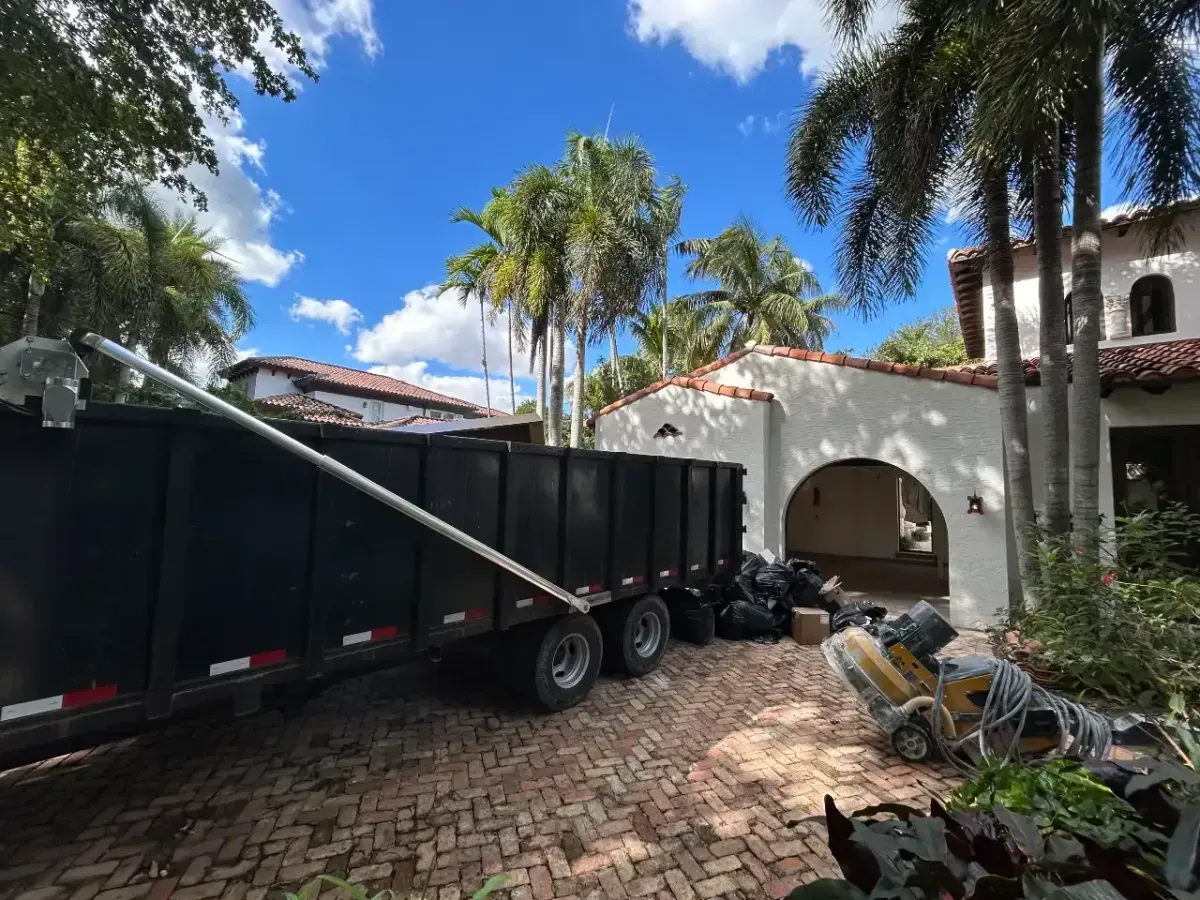 A large black construction dumpster sits on a brick driveway in front of a white house with palm trees under a blue sky.