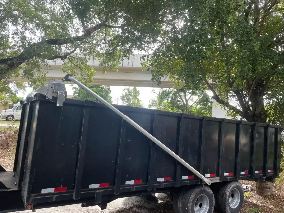 A black dump trailer parked under trees, with a diagonal metal tarp arm extended over the open top.