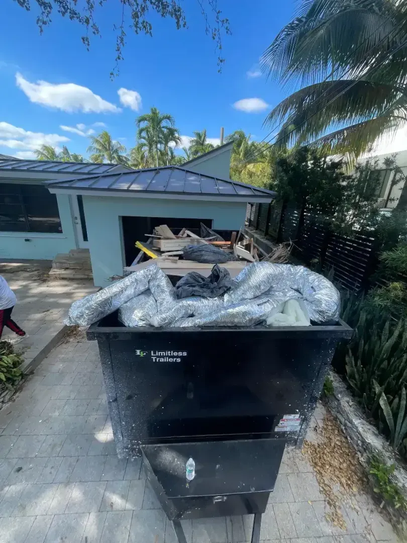 A black dumpster filled with construction debris and metallic air ducts sits in a paved driveway near a house.