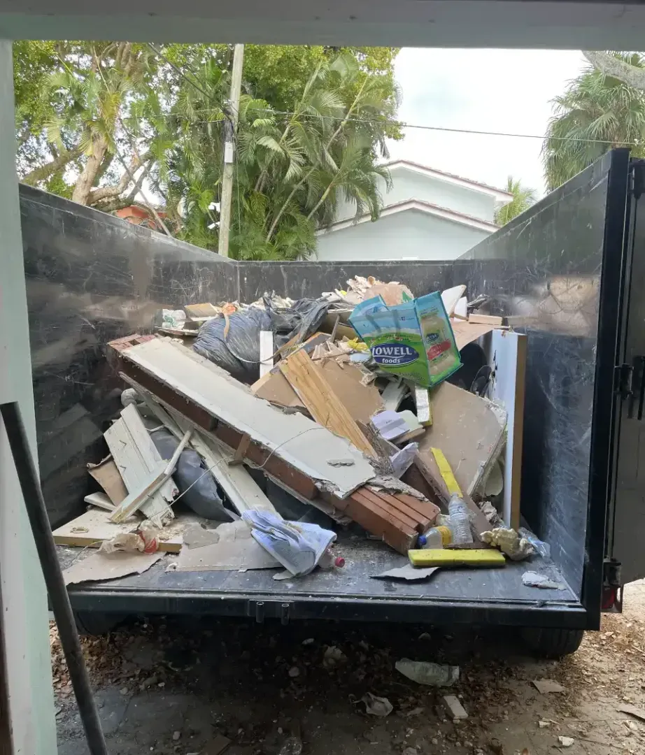 A black metal dumpster filled with construction debris, including wood, cardboard, and trash, sits outdoors.