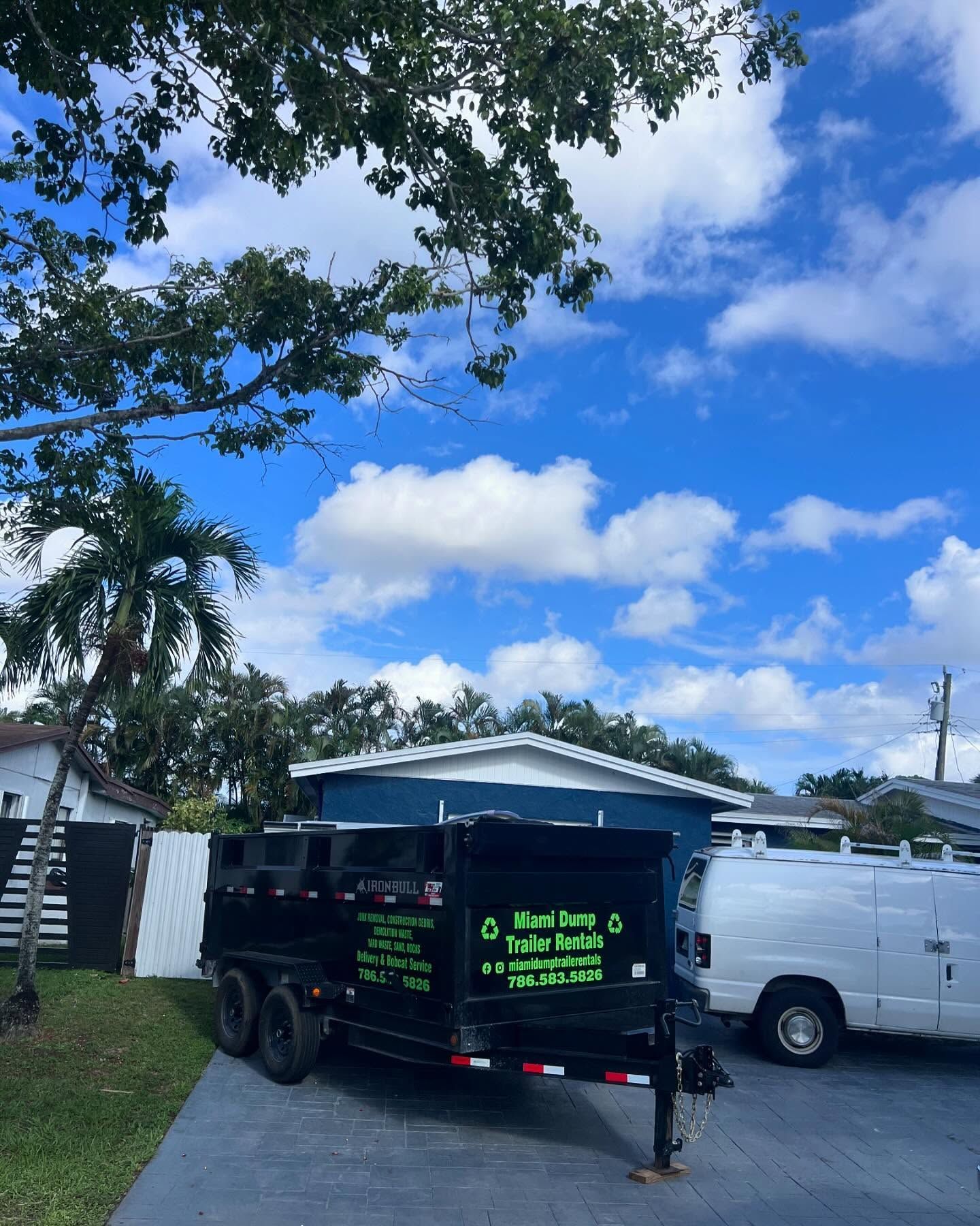 A black dump trailer parked on a residential driveway next to a white van under a blue sky with clouds.