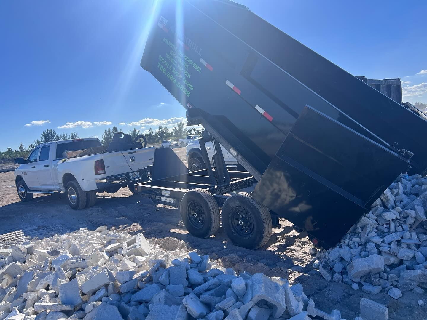 A white pickup truck hauls a black dump trailer in an outdoor lot, tilted to deposit a pile of concrete blocks.