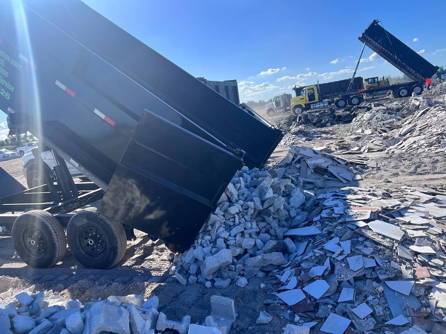Dump trucks are unloading piles of concrete and construction debris at a landfill site under a bright blue sky.
