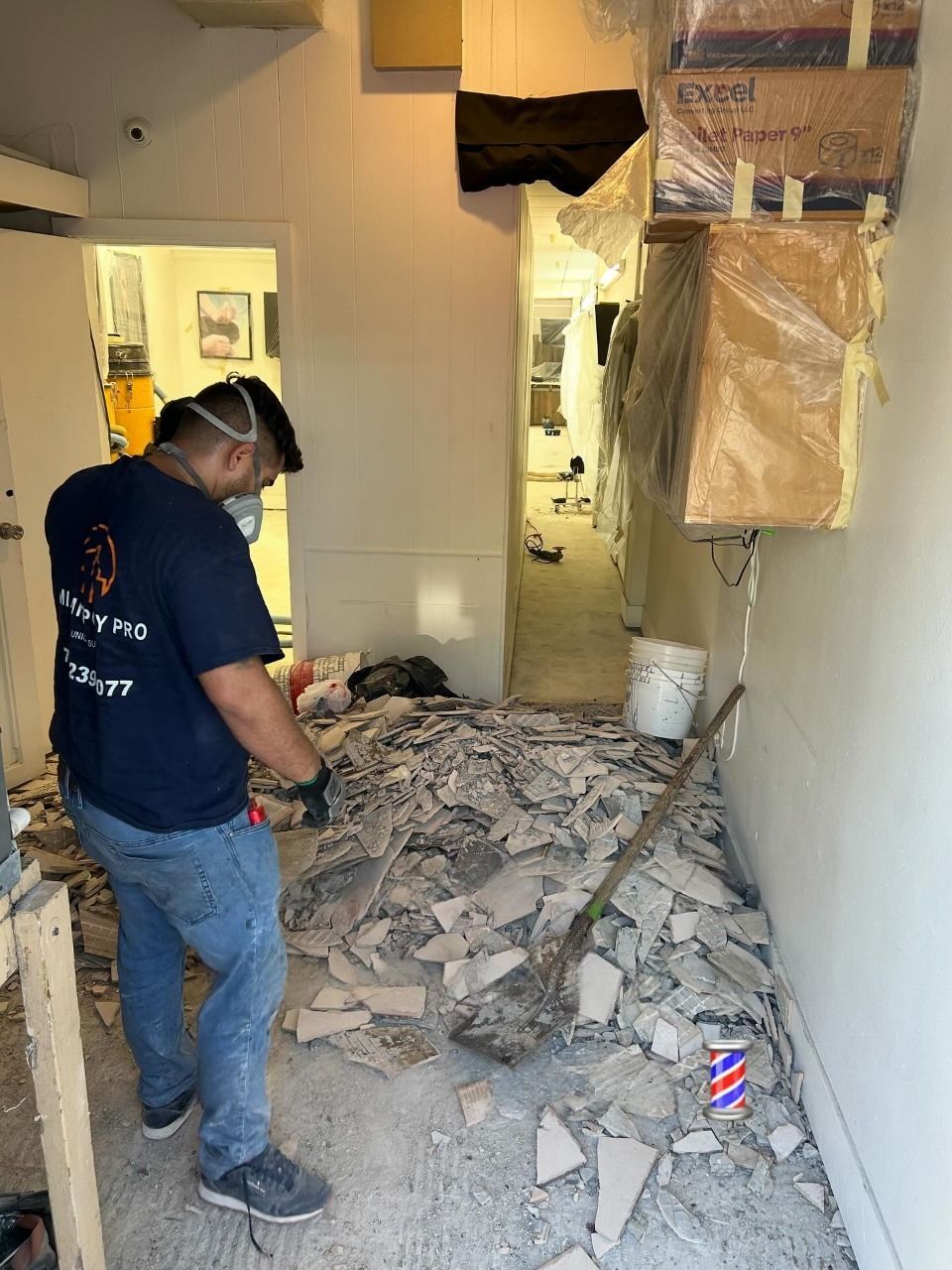 A person in a work shirt and mask cleans up construction debris on a floor with a shovel in a building interior.