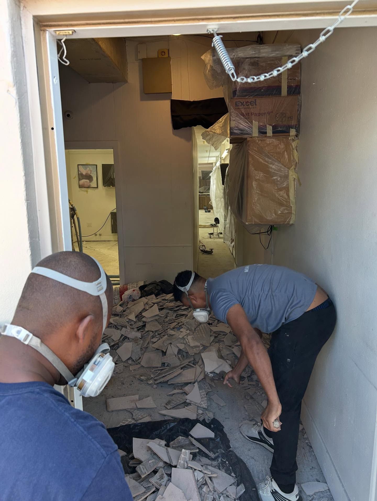 Two people wearing dust masks work inside a small, debris-filled room with boxes stacked on the wall.