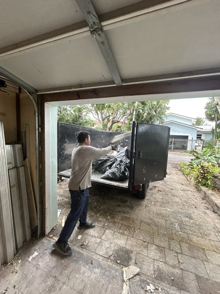 A person throws a black trash bag into a dumpster trailer parked on a concrete driveway outside a garage.
