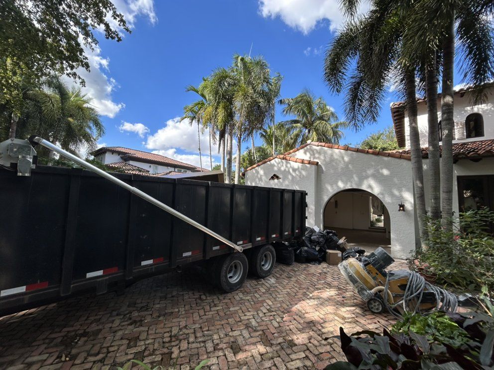 A black construction dumpster parked on a brick driveway in front of a white house with palm trees under a blue sky.