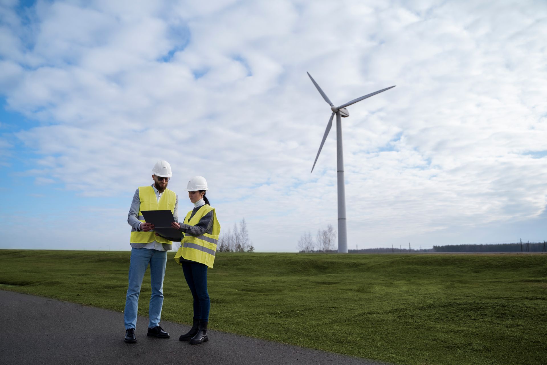 Two people in safety vests and hard hats look at a laptop near a wind turbine.