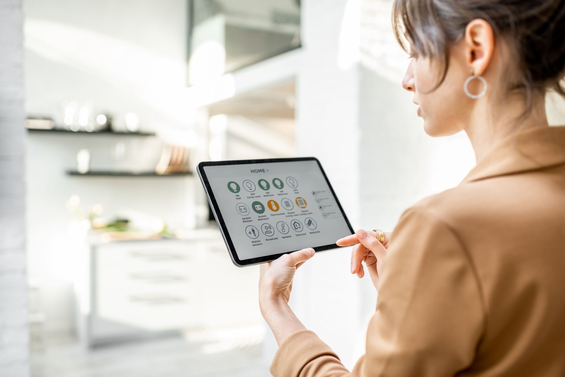 Woman in a tan blazer holding a tablet with a smart home control interface, kitchen in background.