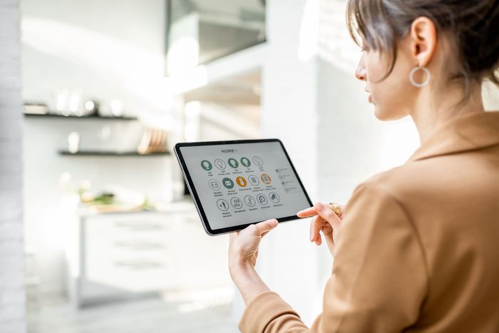 Woman in a tan blazer holding a tablet with a smart home control interface, kitchen in background.