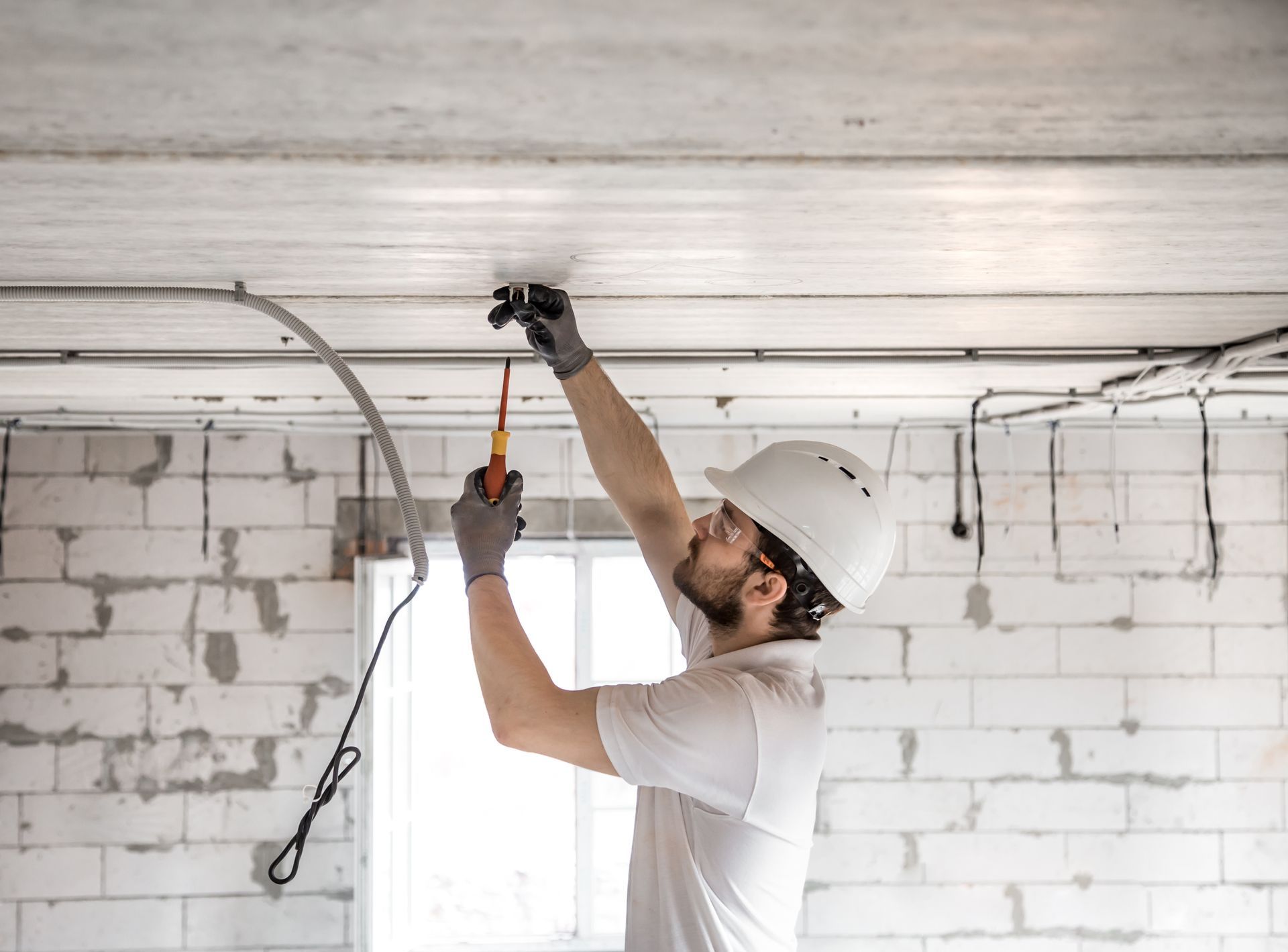 Electrician in white hard hat and gloves working on ceiling wiring. Interior brick construction.