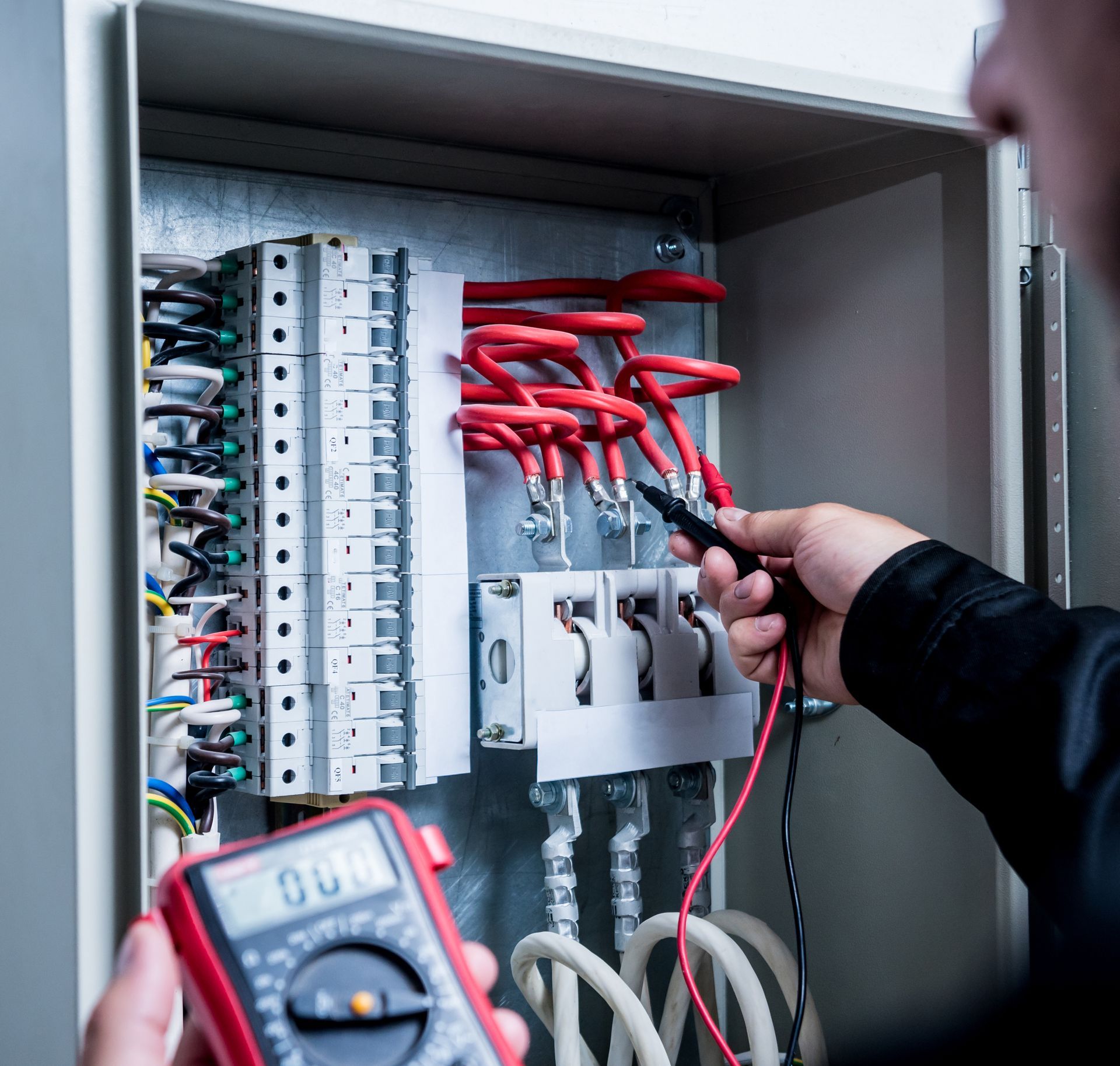 Electrician testing wiring in an open electrical panel with red cables and a multimeter