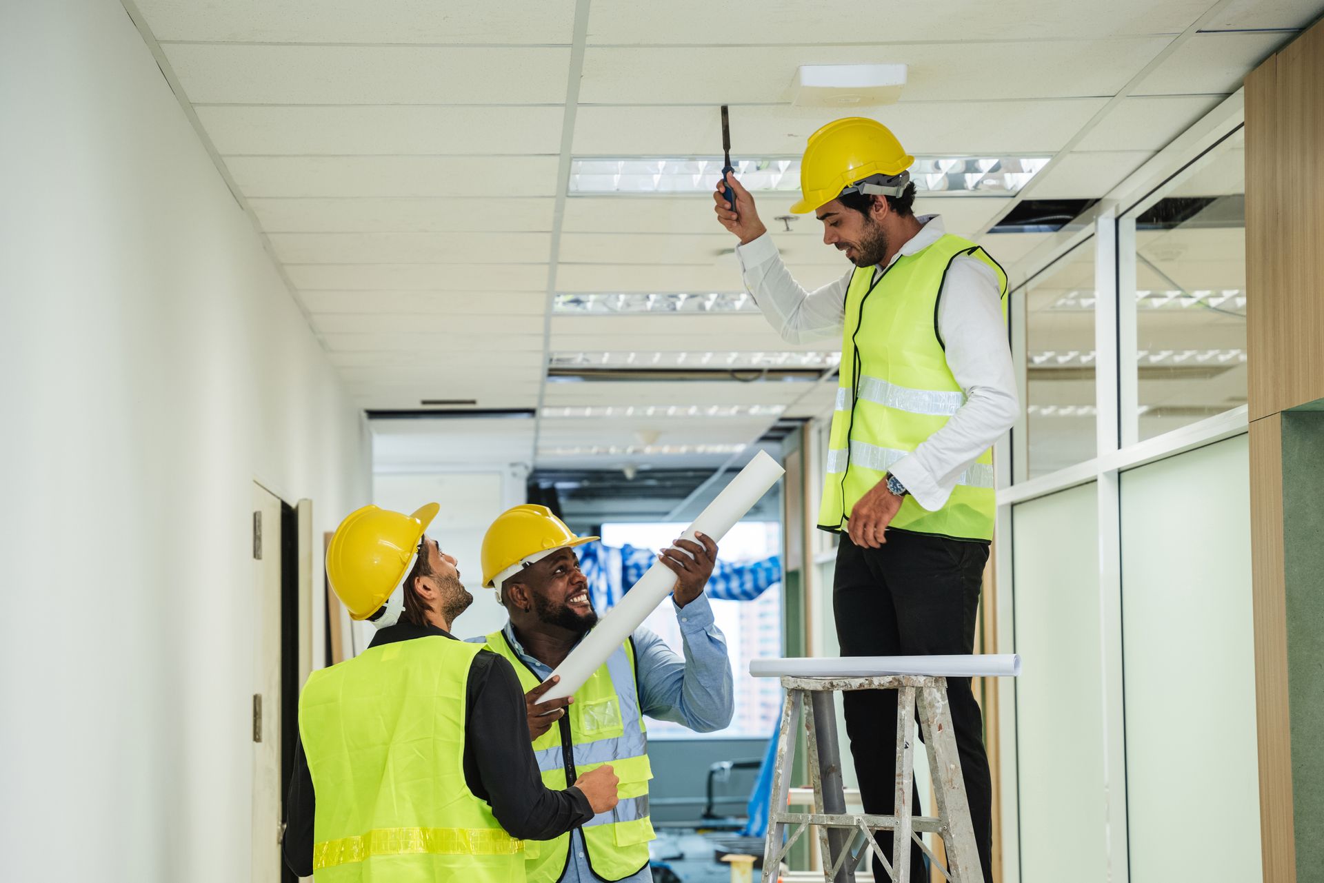 Construction workers inspecting ceiling lights in an office hallway; one on a ladder.