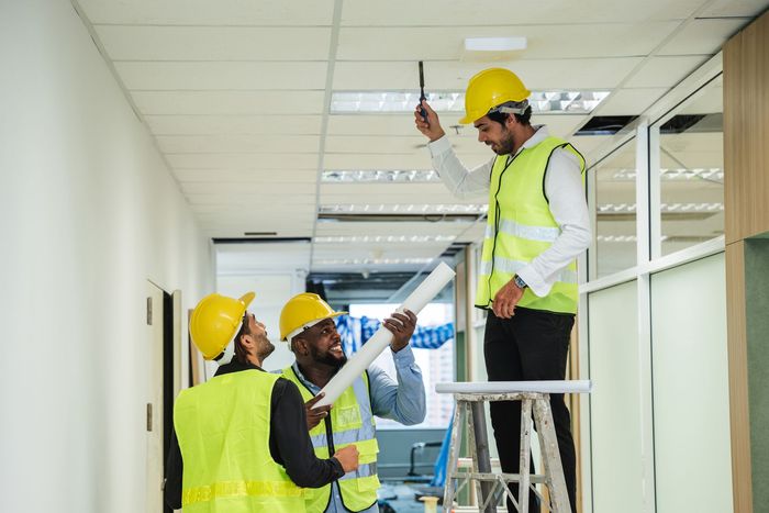 Construction workers inspecting ceiling lights in an office hallway; one on a ladder.