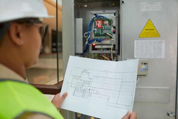 Technician inspecting electrical panel with schematic, wearing a hard hat and safety vest.