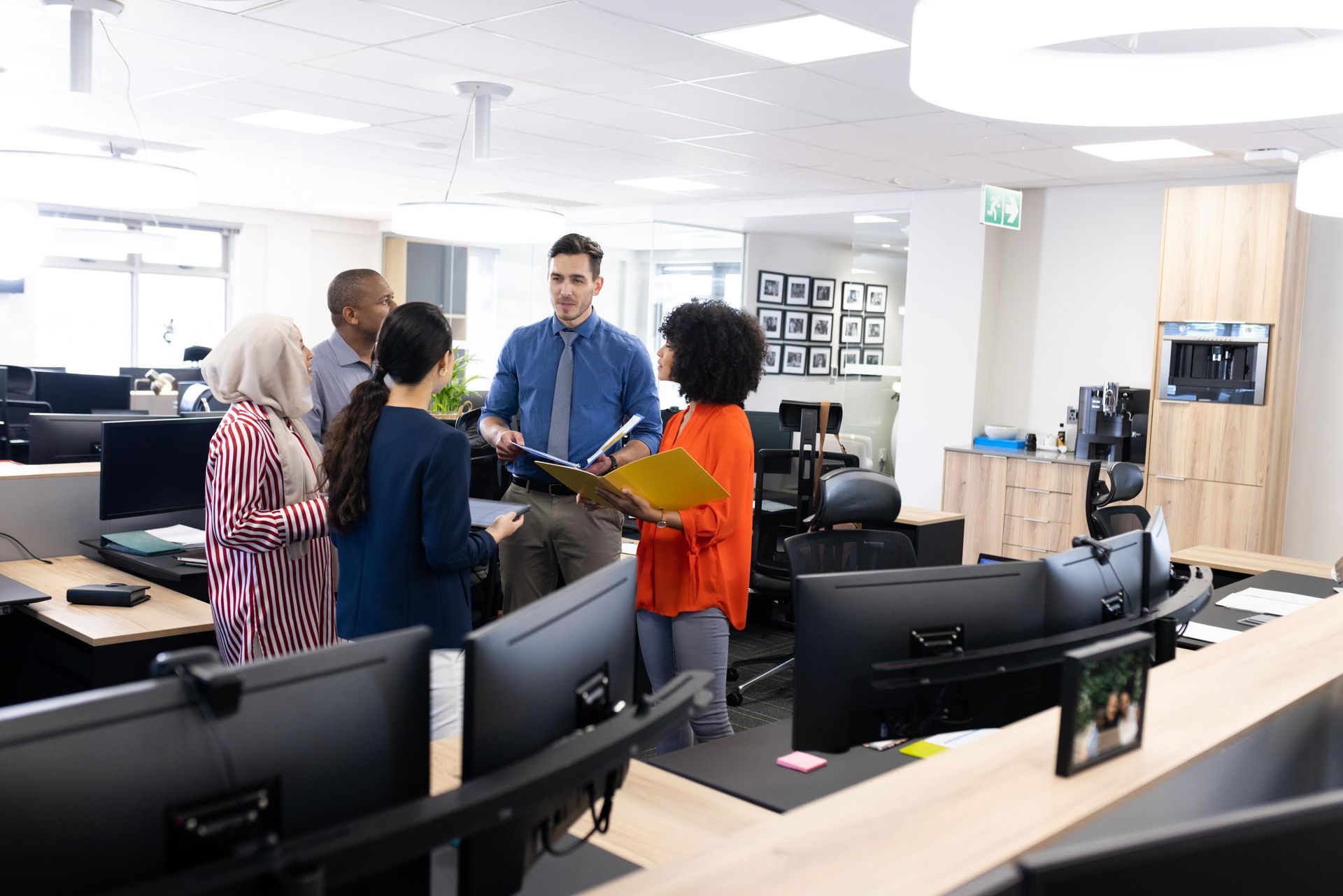 Group of people in an office discussing documents; bright, open space with computers.